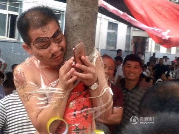 Chứng kiến sự việc lạ lùng, không ít người dùng điện thoại quay phim và chụp lại khoảnh khắc "khó đỡ" này. Chú rể chỉ còn chiếc quần đùi trên người, mặt nhem nhuốc mực đen rồi bị treo lên cột điện và bị dính chặt bằng băng keo.