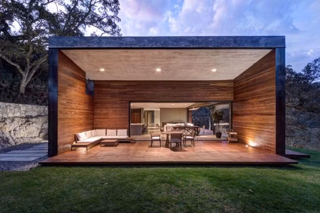 Phòng khách cạnh một đầu ngôi nhà hình cánh bướm nổi bật, lung linh huyền ảo so với cảnh quan thiên nhiên xung quanh lúc gần tối.