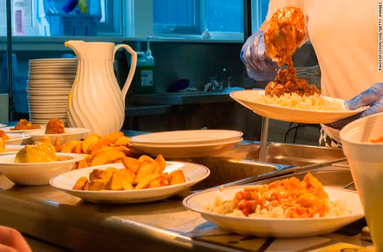 Một người phụ nữ chuẩn bị đồ ăn cho học sinh tại căng tin của trường ở xứ Wales. Thực đơn của bữa ăn bao gồm khoai tây, pizza,...