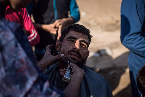 Người đàn ông đang cạo râu, cắt tóc cho một người tị nạn mới đến khu trại Hasansham ở ngoại ô Mosul. (Nguồn ảnh: Al Jazeera).