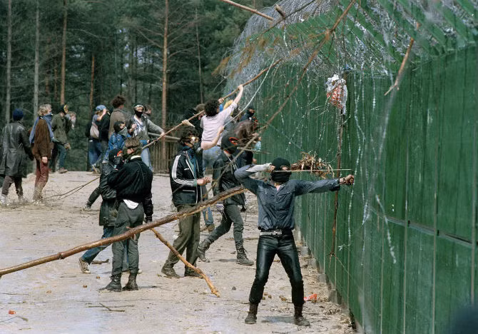 Một người biểu tình đeo mặt nạ dùng súng cao su bắn vào cảnh sát chống bạo động phía sau hàng rào trong một cuộc biểu tình bạo lực tại khu nhà máy tái chế chất thải hạt nhân. Khoảng 10 nghìn người biểu tình phản đối việc sử dụng năng lượng hạt nhân tại Wackersdorf, Tây Đức, ngày 19/5/1986. Hơn 130 cảnh sát bị thương trong cuộc biểu tình này.