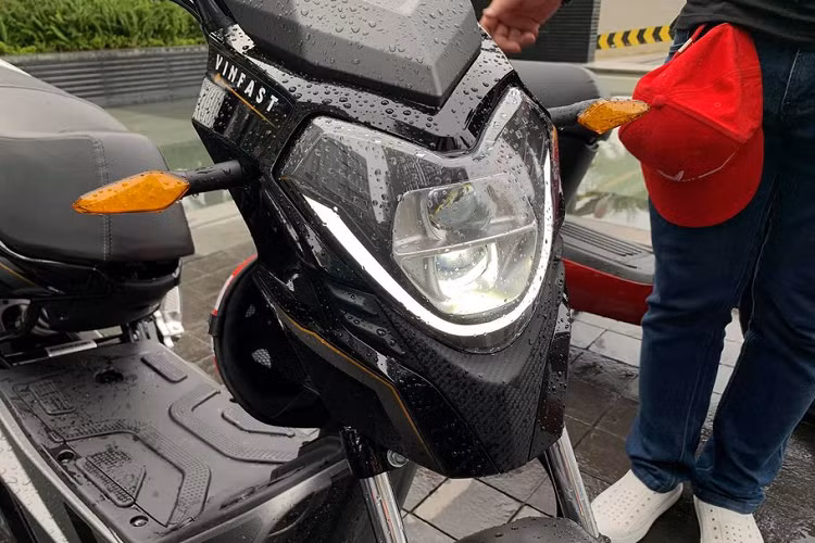 Cận cảnh cụm đèn chiếu sáng LED 2 tầng trên xe máy điện VinFast Impes. Ở phần đầu xe, nổi bật là cụm đèn chiếu sáng dạng LED hình mũi tên với cách bố trí chi tiết 2 tầng pha-cos riêng biệt. Logo VinFast được đặt ở vị trí chắn gió trên to bản và nổi bật.