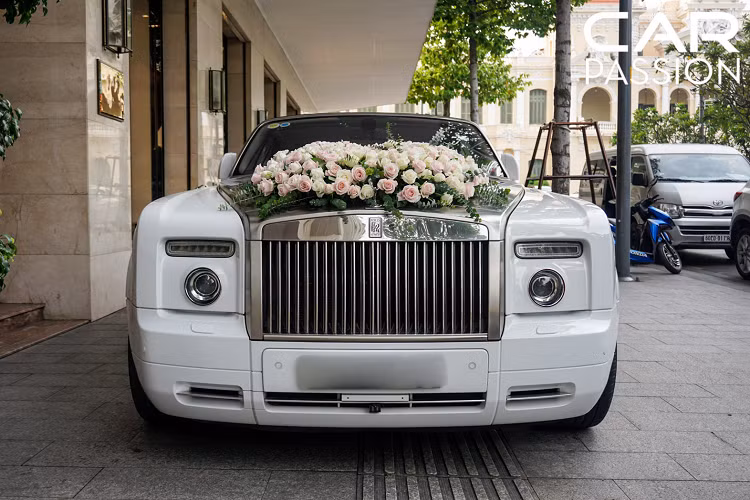 Xuất hiện trong bài viết là một chiếc xe hoa Rolls-Royce Phantom Drophead Coupe với ngoại thất trắng muốt, xen lẫn đó là phần nắp ca-pô được sơn bạc kết hợp cùng phần lưới tản nhiệt “khủng” được mạ chrome sáng loáng. Để phục vụ cho ngày trọng đại, chủ nhân của chiếc xe siêu sang đã trang trí trên phần nắp ca-pô bằng những bông hoa hồng lãng mạn.
