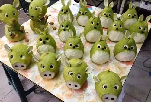 Chú lợn bằng bưởi: Đầu tiên, cắt bỏ phần đầu của quả bưởi. Để quả bưởi nằm ngang rồi ước lượng vị trí cân đối của hai con mắt, khoét phần vỏ xanh đi, để lộ phần cùi trắng. Dùng phần vỏ vừa cắt bỏ để làm tai heo. Sau đó, bạn gắn hạt nhãn vào vị trí hai lỗ mũi và mắt của chú heo.