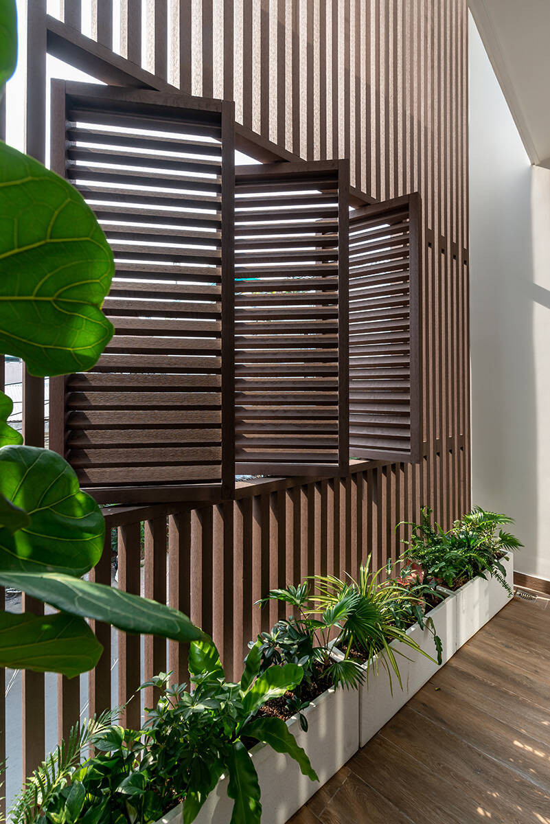 Nhà có mặt tiền thoáng, hướng ra mặt đường nhưng chịu nắng gắt hướng Tây và khói bụi từ xe cộ. Để khắc phục nhược điểm này, nhóm đã bố trí một hệ lam chắn kết hợp cửa sổ lam như một “lớp áo giáp” bao trọn lấy 3 tầng nhà. Giải pháp này vừa che nắng, cản bụi chủ động, vừa giúp không gian trong nhà vẫn đón được ánh sáng, gió trời và giữ gìn sự riêng tư cần có. Phía sau lớp lam chắn là những bồn cây tươi tắn, đóng vai trò như lớp lọc tự nhiên cho nhà.