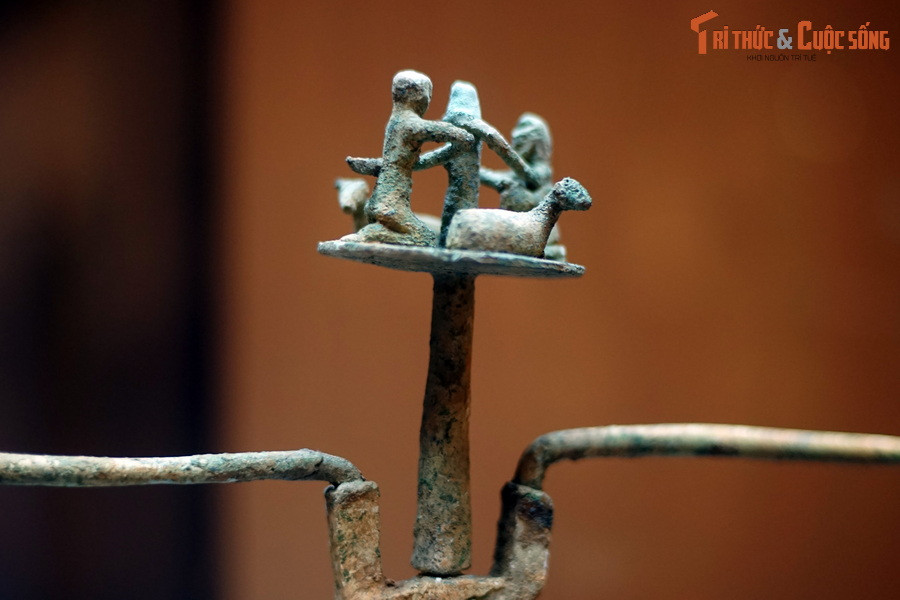 Đỉnh đèn có hình hai người và hai con gia súc, trông giống cừu hoặc dê. Tư thế của hai người giống như đang vận hành một công cụ nào đó.