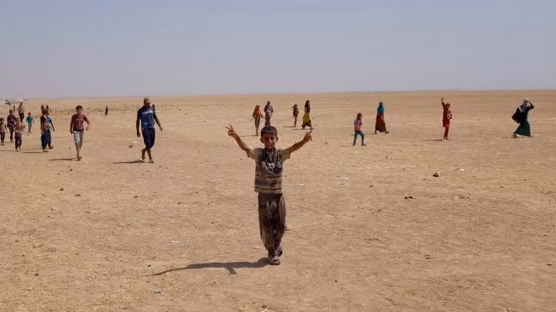 Trước ống kính phóng viên CNN, bé trai đang làm cử chỉ giơ ngón tay lên biểu thị cho sự chiến thắng. Em cùng nhiều người dân khác hiện sống trong điều kiện khắc khổ tại các trại tị nạn bên ngoài thị trấn Qayyarah. Họ phải sống giữa vùng sa mạc cằn khô, thiếu nước uống và hứng chịu những trận nóng bức khắc nghiệt.