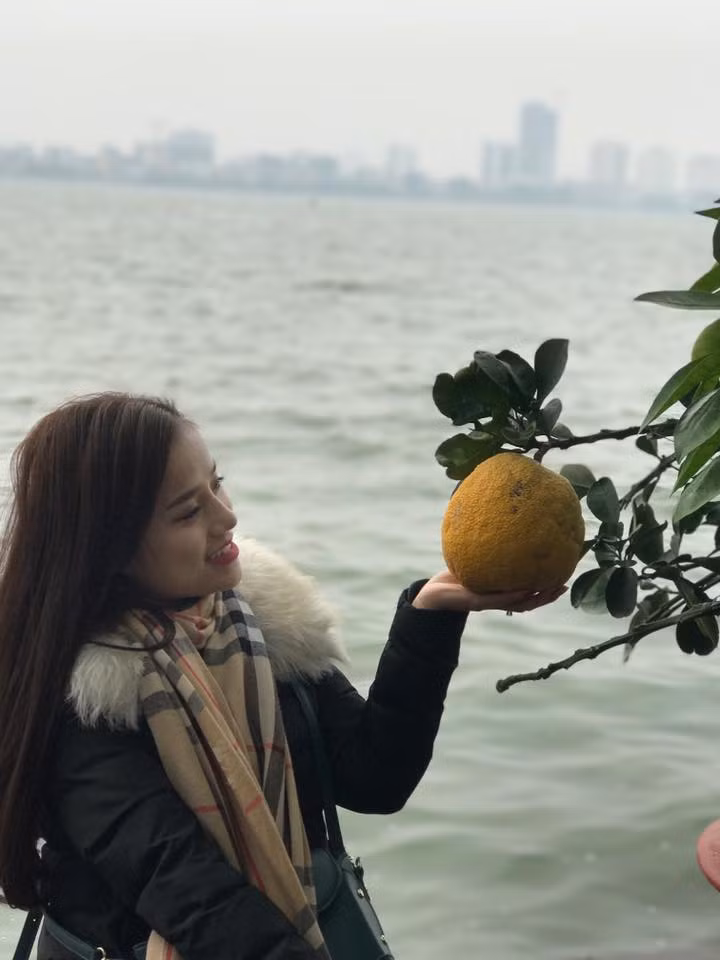 Cô gái xinh đẹp đang nở nụ cười tươi, giơ lòng bàn tay lên nhẹ đỡ lấy quả bưởi để chụp ảnh...