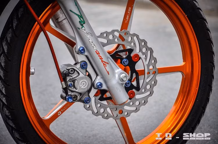 Trong đó, chi tiết nổi bật nhất của bản độ này là cặp mâm Racing Boy 5 cây CNC thể thao, sắc sảo và có màu cam đúng phong cách Repsol. Hệ thống phanh trước của chiếc xe cũng được nâng cấp khá "khủng" với đĩa Braketech và heo Brembo 2 pis đối xứng.