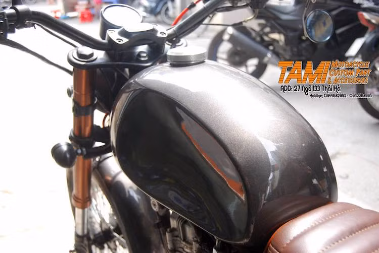 Điểm nhấn nổi bật của bản độ này là bình xăng được gò thủ công tròn trịa với hai bên làm lõm vào, tạo điểm tì cho đầu gối người lái. Nắp xăng bằng nhôm tiện CNC cũng góp phần tăng sự tinh tế cho chiếc xe. Hai bên thân xe là cặp ốp nhựa nguyên bản được sơn màu xám ánh kim như bình xăng thủ công.