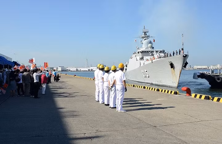 Các sĩ quan Lực lượng Phòng vệ trên biển Nhật Bản (JMSDF) xếp hàng chờ bắt dây neo để cố định tàu Trần Hưng Đạo vào cầu tàu. Phía sau là người dân đang vẫy quốc kỳ Việt Nam và Nhật Bản.