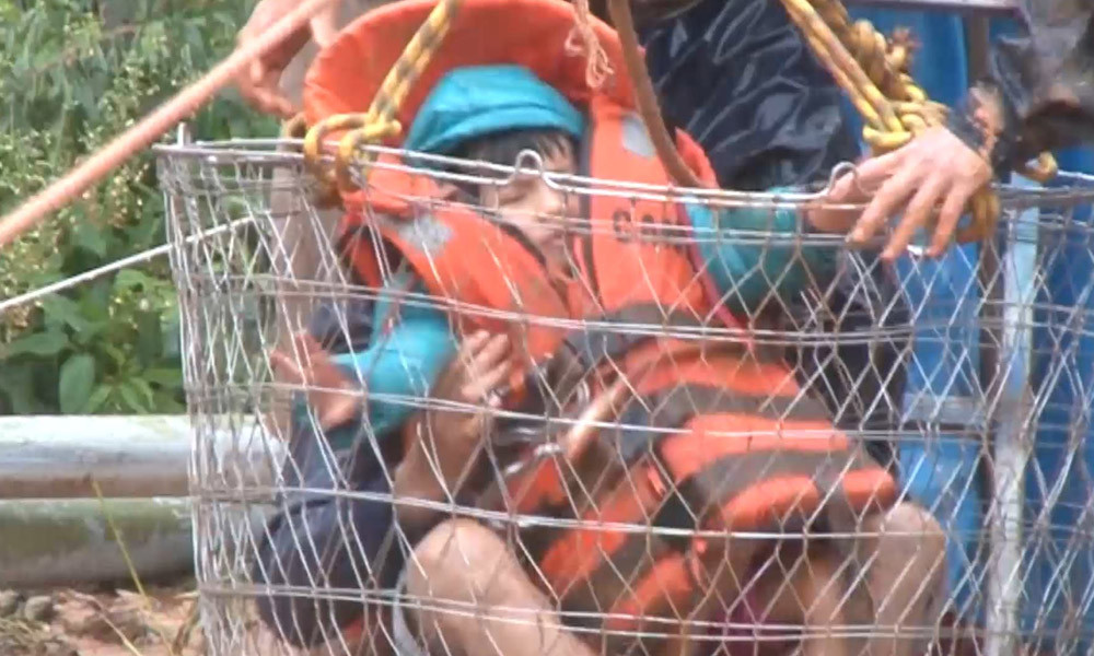 Trong ảnh là một bé trai đã được cho mặc áo phao. Em bé được đặt trong một giỏ sắt, hai tay nắm chặt dây thừng buộc thành giỏ với cáp cứu hộ.