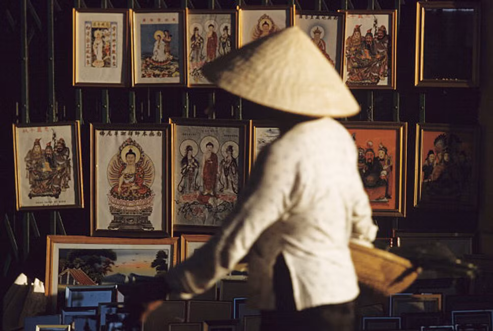 Một người phụ nữ đội nón lá đi qua cảnh xửa xếp treo đầy tranh ảnh tôn giáo của người Hoa ở Chợ Lớn.