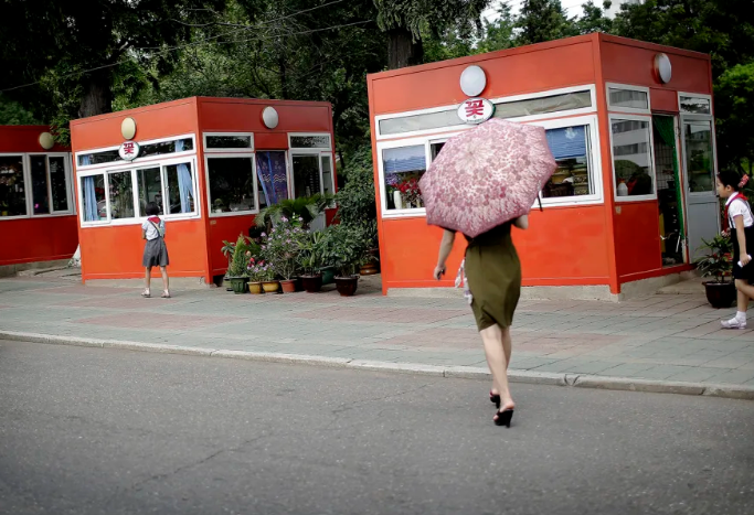Người phụ nữ che ô đi về phía các ki-ốt bán hoa, đồ ăn nhẹ và đồ uống tại Bình Nhưỡng vào ngày 18 tháng 7 năm 2017.