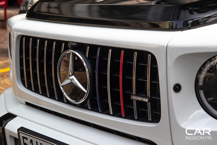 Chiếc Mercedes-AMG G63 này mang màu gốc là màu trắng bóng nhưng đã được phối lại với phong cách hai tông màu quen thuộc trên những mẫu sedan sang trọng. Phần mui xe được dán decal màu đen bóng tương phản, kết hợp với đó chính là decal ba màu đỏ-trắng-xanh mang đậm dấu ấn của chủ sở hữu. Những đường màu này được phối trên các chi tiết như phần cản trước xe, hộp đựng bánh phụ phía sau hay cả họa tiết trên cánh cửa.