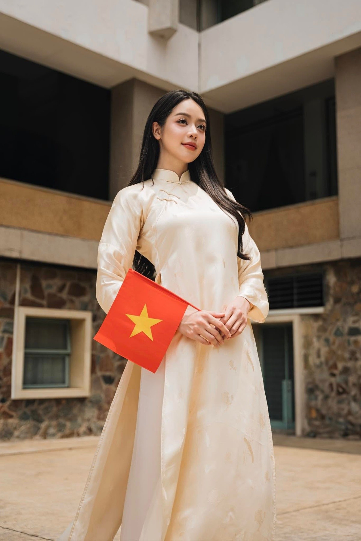 Trong ảnh, đương kim Hoa hậu Quốc tế 2024 diện thiết kế áo dài màu nâu vàng nhạt, tạo dáng duyên dáng. Cô còn cầm lá cờ Tổ quốc bé trên tay.