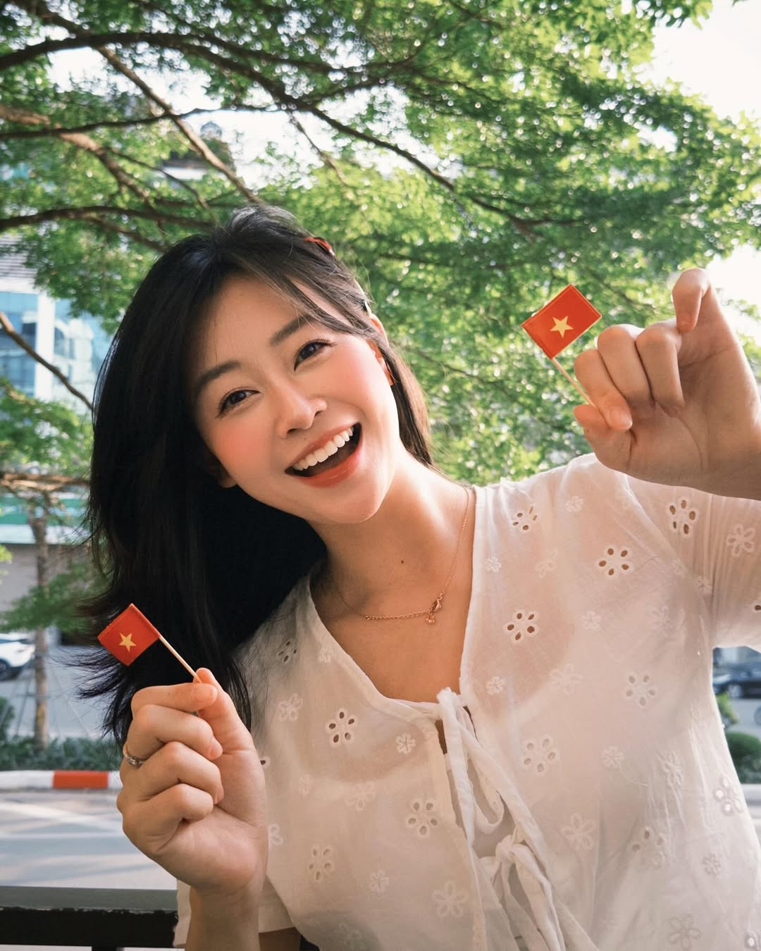 Lê Chi lựa chọn trang phục với tông màu pastel nhẹ nhàng, như áo cardigan vàng nhạt kết hợp với chân váy trắng dài, tạo nên sự hài hòa với sắc đỏ rực rỡ của cờ Tổ quốc.