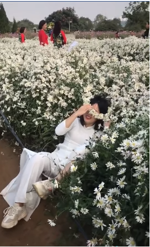 Cô gái trẻ trong bộ áo dài trắng, đầu đội thêm chiếc vòng bện từ hoa cúc đang cố gắng dang tay nghiêng người để tạo dáng chụp ảnh.