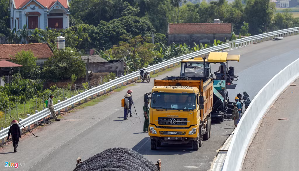 Công nhân sử dụng máy trải bê tông nhựa nóng, phía trước có xe tiếp nguyên liệu, phía sau có xe lu thảm. 
