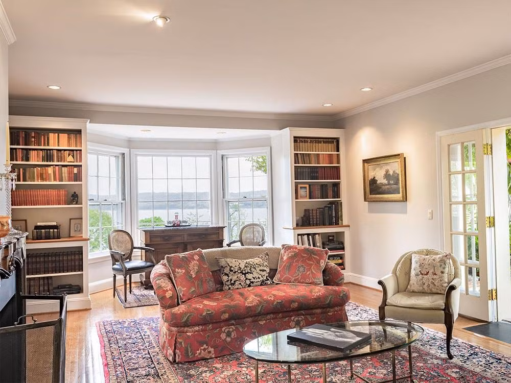 Phòng khách đầy trang nhã với lò sưởi, tủ sách cao, bộ sofa phong cách cổ điển và ô cửa sổ rộng lớn phóng ra quang cảnh bờ sông.