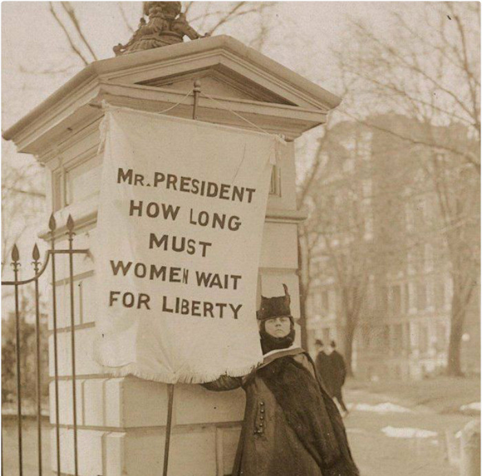 Một người phụ nữ biểu tình trước Nhà Trắng năm 1917 với dòng chữ: "Ngài Tổng thống, phụ nữ phải chờ đợi bao lâu cho tự do".