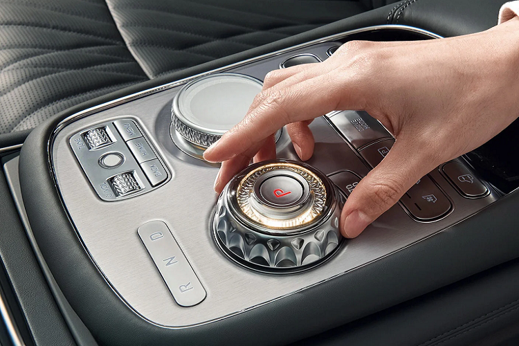 Hai màn hình trên Genesis G90 2023 hạng sang được ngăn cách nhau bởi 1 mảng trang trí cùng màu tap lô. Các khe điều hòa được thu hẹp và ẩn mình ở khu vực này, trong khi cụm điều khiển nằm nhỏ gọn bên dưới màn hình. Giữa xe, ngang với bệ tỳ tay là cụm cần số và núm điều chỉnh chế độ lái cao cấp màu bạc.