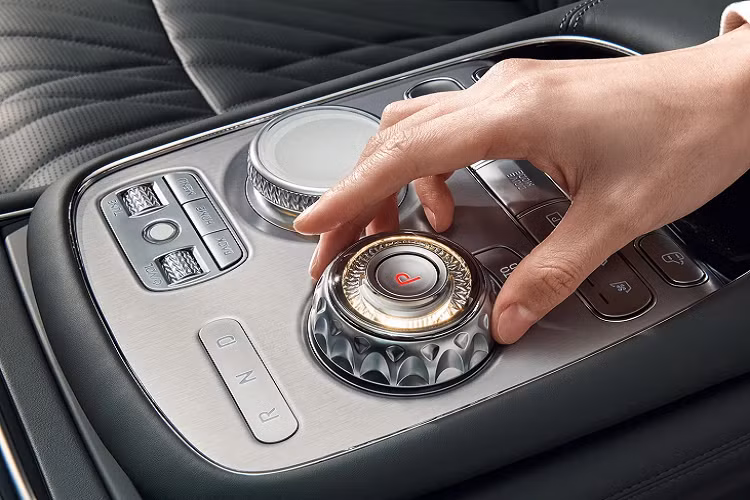 Hai màn hình trên Genesis G90 2023 hạng sang được ngăn cách nhau bởi 1 mảng trang trí cùng màu tap lô. Các khe điều hòa được thu hẹp và ẩn mình ở khu vực này, trong khi cụm điều khiển nằm nhỏ gọn bên dưới màn hình. Giữa xe, ngang với bệ tỳ tay là cụm cần số và núm điều chỉnh chế độ lái cao cấp màu bạc.