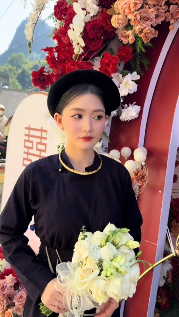 Cô dâu sở hữu làn da trắng, gương mặt thanh tú, nụ cười hiền dịu, trang điểm vừa phải, cầm bó hoa cưới đứng ở cổng chào đón khách mời.