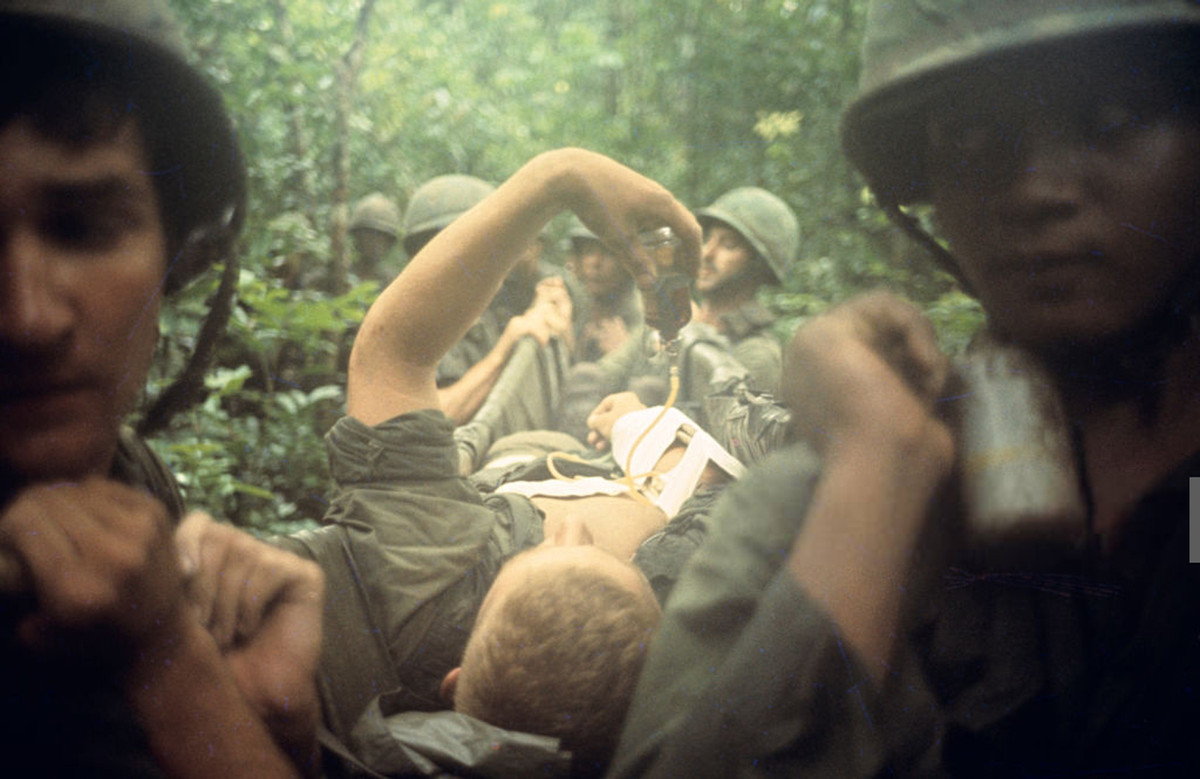 Lính Mỹ bị thương do một vụ nổ được đồng đội nẹp cánh tay và cáng ra vị trí trực thăng cứu hộ. Tay trái của người lính này cầm ống truyền thuốc. Ảnh: Jean-Claude Sauer.
