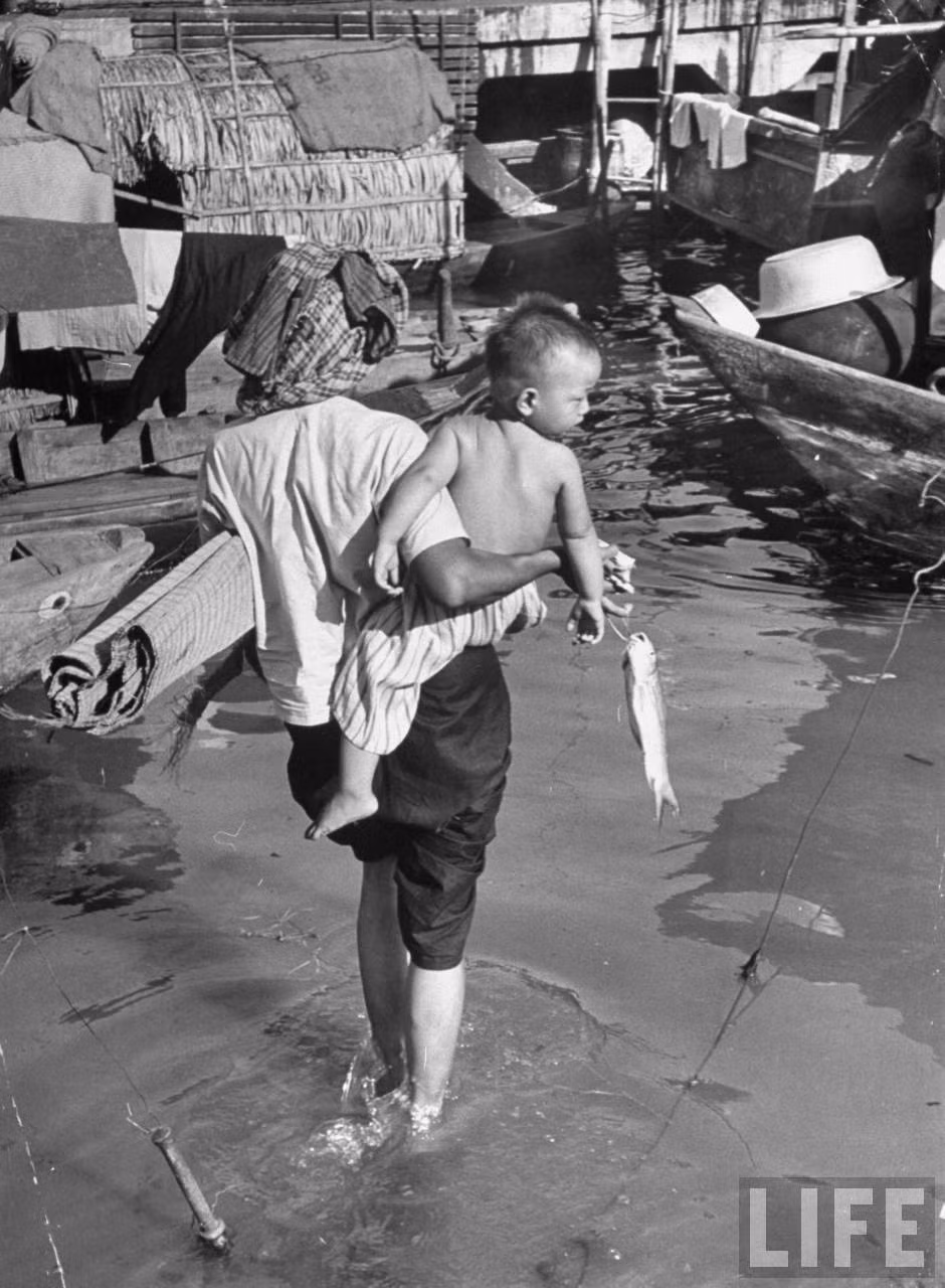 Bà mẹ bế con nhỏ về thuyền, tay cầm con cá chuẩn bị được chế biến cho bữa ăn. Ảnh: Carl Mydans/ Life.