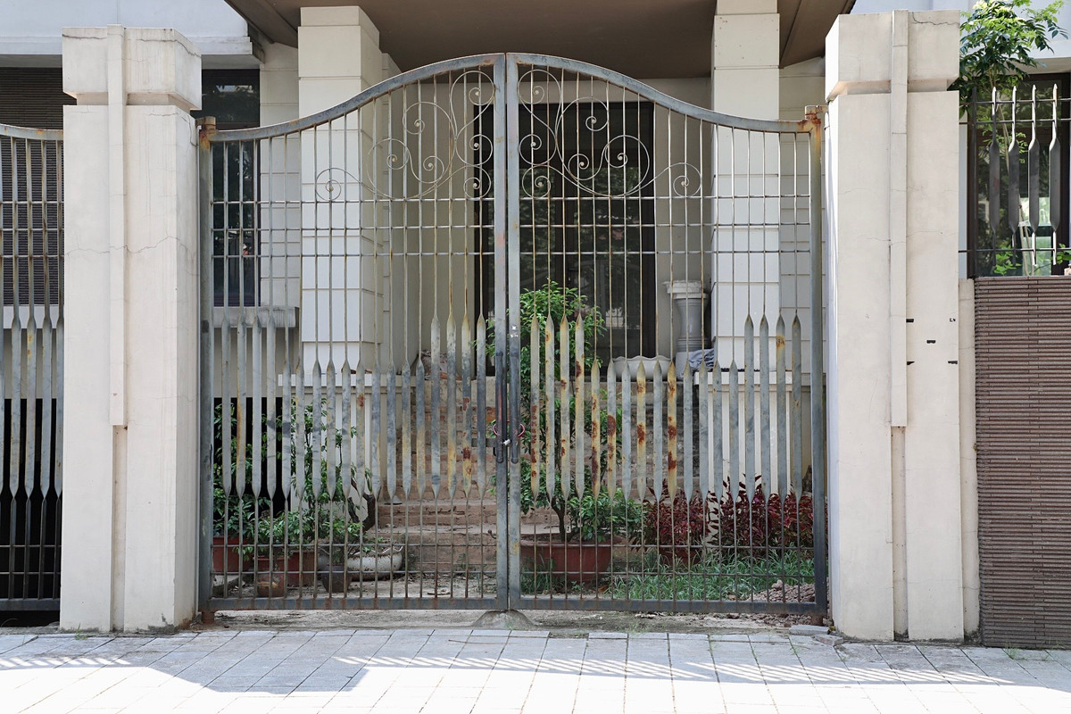 Cổng sắt hoen rỉ, phía bên trong sân cỏ cây mọc um tùm.