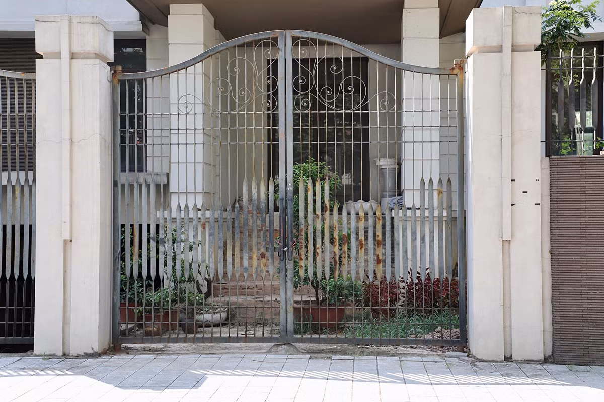 Cổng sắt hoen rỉ, phía bên trong sân cỏ cây mọc um tùm.