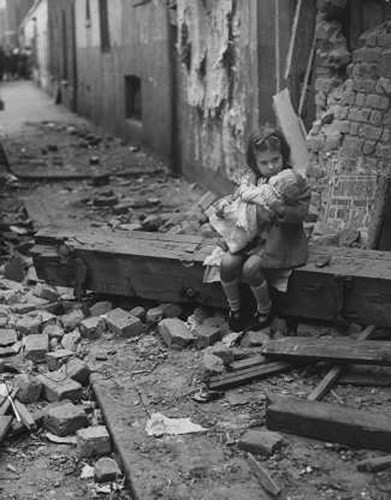 Một bé gái ngồi ôm búp bê trước ngôi nhà bị ném bom.