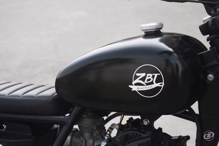 Trong khi đó, bình xăng nguyên bản của GN250 cũng đã được chỉnh sửa lại một chút, sơn màu đen bóng và kết thúc với phần nắp nhôm CNC do chính DuongDoan Design tiện bằng máy. Yên của chiếc xe khá mỏng và có vân ngang dạng yên "bánh mì, chạy thẳng theo khung phụ theo đúng phong cách brat.