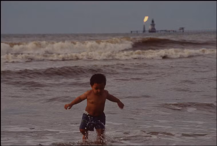 Một em bé chơi đùa trên bờ biển, phía xa là giàn khoan của Brunei. Ảnh: Gueorgui Pinkhassov.