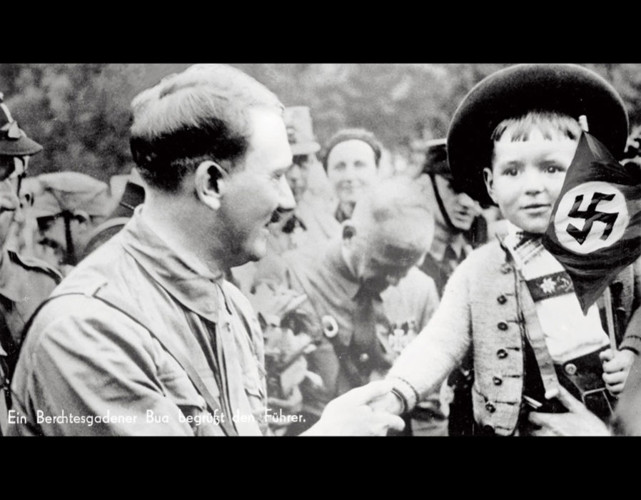 Trùm phát xít bắt tay với một cậu bé đang giơ lá cờ chữ thập ngược - biểu tượng của Đức Quốc xã.