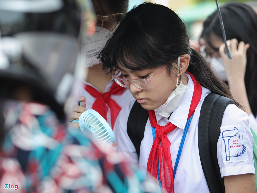 Một số học sinh trang bị quạt cầm tay để tránh nóng. Các em đứng chen chúc trong bóng râm trước cổng trường.