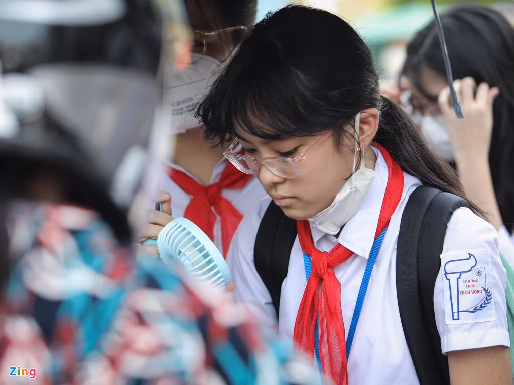 Một số học sinh trang bị quạt cầm tay để tránh nóng. Các em đứng chen chúc trong bóng râm trước cổng trường.