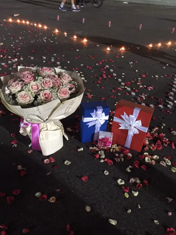 Cùng với dòng chữ "LOVE", ở chính giữa trái tim lớn được xếp bằng nến có đặt một bó hoa hồng và 3 hộp quả từ lớn, nhỏ. Đây là những món quà mà chàng trai trẻ tên Hoàng Anh - chủ nhân của màn cầu hôn này dành tặng cho bạn gái của mình.