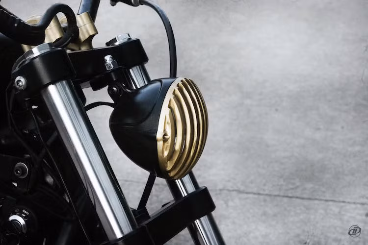 Ở bản độ này, chiếc 883 dã được sơn lại trong màu đen mờ, kết hợp cùng các chi tiết đồng màu vàng tạo nên sự tương phản độc đáo. Chi tiết đầu tiên bằng đồng CNC đó là phần lưới dọc chắn chắn chóa pha ở phía trước chiếc xe.