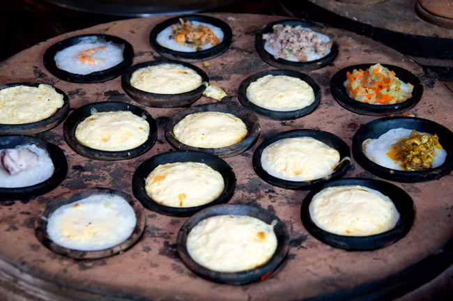 Bánh căn: Bánh căn gần giống bánh khọt, nhưng làm bằng bột gạo pha nếp, trên lò than hồng là khuôn bánh bằng đất nung có đến cả chục khuôn nhỏ giống như khuôn bánh khọt. Người đổ bánh có kinh nghiệm làm sao bánh khi ra khỏi khuôn phải vừa giòn, vừa xốp lại vừa dẻo, vỏ bánh phải có màu trắng vàng nhưng không bị cháy khét. Bánh căn được nướng chín bằng khuôn đất nung nên ăn cũng không mau ngán. Ảnh: Dulich24.