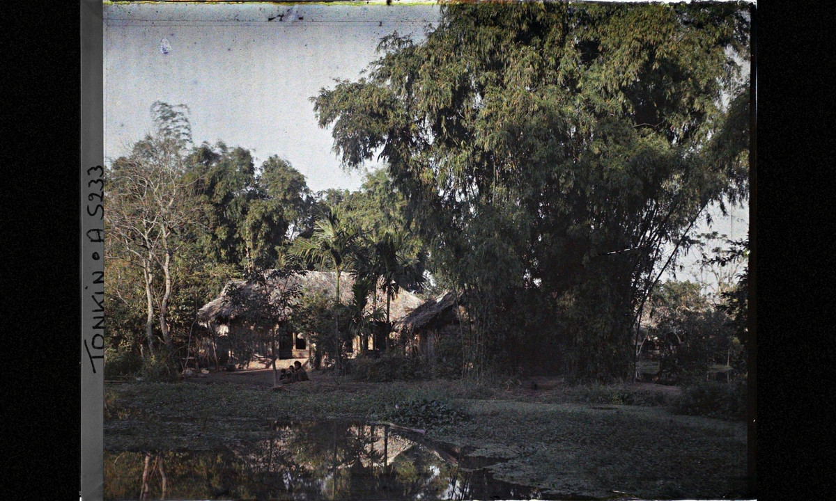 Ao nước, bụi tre và những ngôi nhà lá. Người phụ nữ và hai đứa ngồi bên bờ ao. (Nguồn: Léon Busy) 