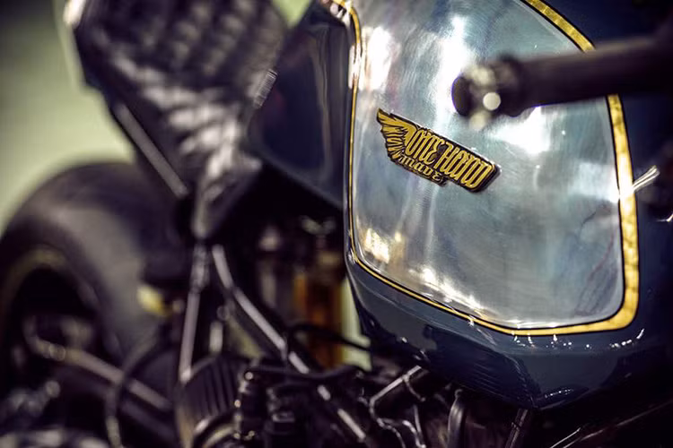 Bình xăng được gò lại thủ công bằng chất liệu nhôm, trên bề mặt không sơn được đánh bóng hiển thị logo tên xưởng độ Onehandmade. Dàn chân trước sử dụng phuộc hành trình của hãng Ohlins.