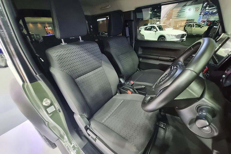 Bên trong xe, Suzuki Jimny 1.5 AT 4WD 2023 tại Thái Lan chỉ có ghế bọc nỉ màu đen, ghế lái chỉnh tay 6 hướng, ghế phụ lái chỉnh tay 4 hướng, ghế sau gập 50:50, vô lăng 3 chấu bọc da, chỉnh 2 hướng, tích hợp phím chỉnh âm lượng/đàm thoại rảnh tay và cửa sổ 1 chạm chống kẹt bên ghế lái.