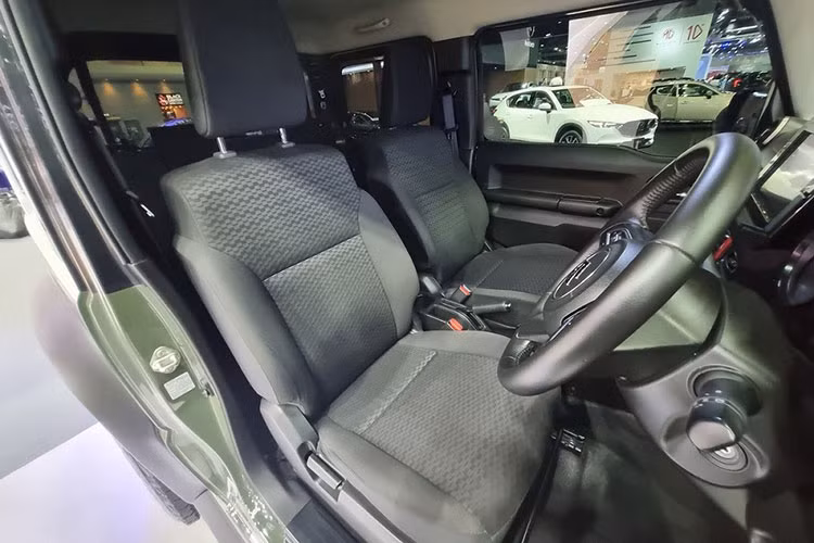 Bên trong xe, Suzuki Jimny 1.5 AT 4WD 2023 tại Thái Lan chỉ có ghế bọc nỉ màu đen, ghế lái chỉnh tay 6 hướng, ghế phụ lái chỉnh tay 4 hướng, ghế sau gập 50:50, vô lăng 3 chấu bọc da, chỉnh 2 hướng, tích hợp phím chỉnh âm lượng/đàm thoại rảnh tay và cửa sổ 1 chạm chống kẹt bên ghế lái.