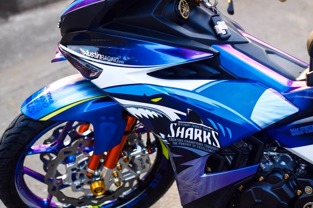 Màu sơn của chiếc xe côn tay cỡ nhỏ đình đám nhà Yamaha cũng đã được chủ nhân chuyển xanh dương - tím, trên từng khoảng màu xe đều được nhấn nhá thêm các đường nét màu vàng, trắng sao cho phù hợp với màu trên dàn đồ chơi độ đầy cá tính trang bị trên xe.