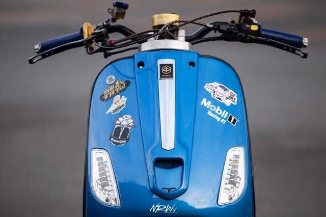 Đầu đèn của mẫu xe tay ga Vespa LX đã được tháo bỏ, thay vào đó là cụm ghi đông trần với trợ lực trung tâm Scott. Phần yếm trước vẫn giữ nguyên nhưng được chủ nhân dán thêm các hình dáng tạo điểm nhấn và nét riêng.