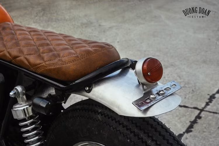 Ở phía sau, bộ khung phụ của EN150A được chế lại, phù hợp với chiếc yên bọc da, chần chỉ màu nâu đầy "phong trần. Cả 2 dè trước và sau xe đều đã được Duong Doan Design gò từ nhôm và để nguyên màu sắc nguyên bản của loại vật liệu này, không phủ màu.