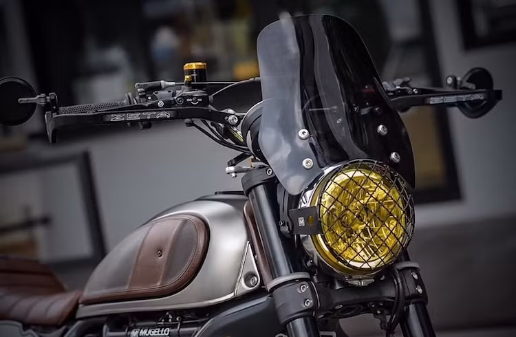 Ở phía trước, dàn đầu nguyên bản của Scrambler đã được độ lại rất "bụi" với lưới chắn pha mắt cáo chính hãng Ducati cùng chóa đèn dán màu vàng. Nằm phía trên cụm đèn pha của xe là kính chắn gió lớn, hạn chế gió tạt ở tốc độ cao.