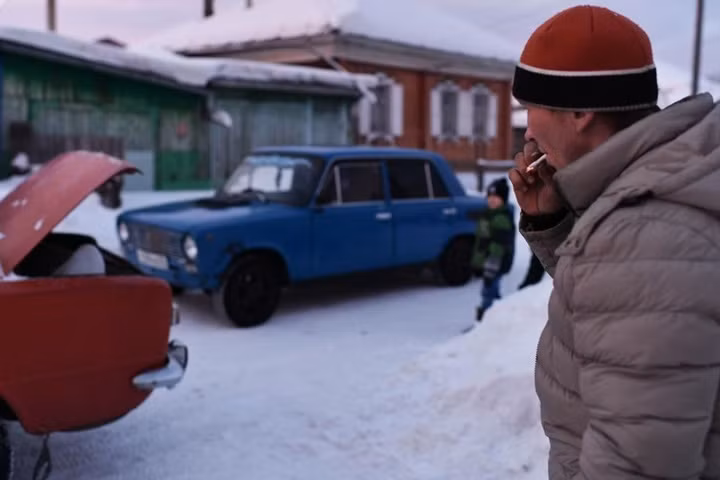 Một nam giới mặc áo phao đội mũ len hút thuốc bên ô tô con ở Tara.